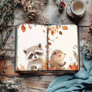 Puede incluir: Un diario abierto con ilustraciones de un mapache y un erizo rodeados de hojas de otoño. El diario está sobre una superficie de madera rústica, con una taza de té y detalles florales en el fondo.
