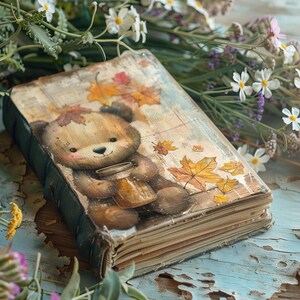 Peut inclure: Un journal de style vintage avec une couverture en cuir marron et une illustration peinte d'un ours tenant une tasse. Le journal est entouré de fleurs blanches et violettes.