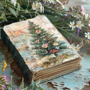 Puede incluir: Un diario vintage de tapa azul, encuadernado en cuero, con una imagen pintada de un árbol de Navidad en la portada. El diario está rodeado de flores blancas y moradas.