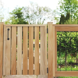 Könnte beinhalten: Ein Holzzaun mit einem schwarzen Griff und Scharnieren. Das Tor besteht aus vertikalen Holzplanken und hat einen Drahtgitterabschnitt. Im Hintergrund sind Bäume und Grün zu sehen.