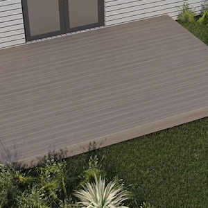 Puede incluir: Una terraza rectangular de madera marrón claro con tablones horizontales. La terraza está rodeada de hierba verde y varias plantas. La terraza está situada frente a una casa blanca con una puerta con marco oscuro.