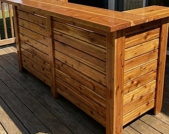 Puede incluir: Una barra de exterior de madera con acabado natural. La barra está construida con tablones horizontales y cuenta con una encimera y una sección inferior. La barra está diseñada para uso en exteriores y está hecha de madera.