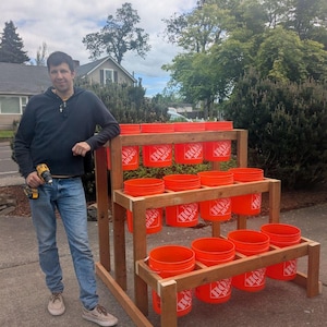 May include: A wooden tiered planter filled with orange buckets. The planter is constructed from natural wood and holds multiple buckets, creating a multi-level gardening system. The buckets are bright orange and labeled "Home Depot".