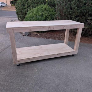 May include: A rectangular workbench constructed from light-colored unfinished wood. The structure features a flat top surface, a lower shelf, and four vertical support legs. The entire unit is mounted on small black caster wheels for mobility.