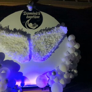 May include: A white backdrop with a round, white sign that says "Dominic's Baptism" with a baby sleeping inside a crescent moon. The sign is decorated with white feathery wings. The backdrop is decorated with white and silver balloons.