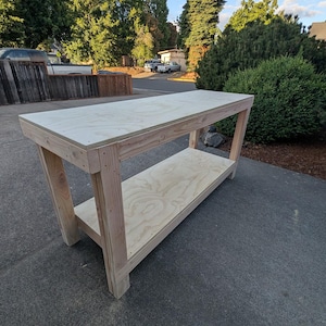May include: A rectangular workbench constructed from light-colored plywood and dimensional lumber. The structure features a flat, smooth top surface and a lower storage shelf supported by four sturdy vertical legs, showcasing natural wood grain.