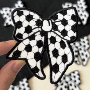 May include: A fabric bow patch featuring a black and white soccer ball design. The bow has a black outline. Other similar patches are visible in the background.