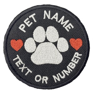 May include: A round, black embroidered patch with white text that reads "PET NAME" and "TEXT OR NUMBER". A white paw print is in the center, flanked by two red hearts. The patch is designed for pets.