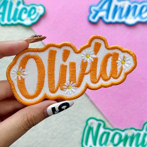 May include: An embroidered patch with the name "Olivia" in orange script on a white background, outlined in orange. Small white and yellow daisy accents are scattered around the name. Other name patches are visible in the background.