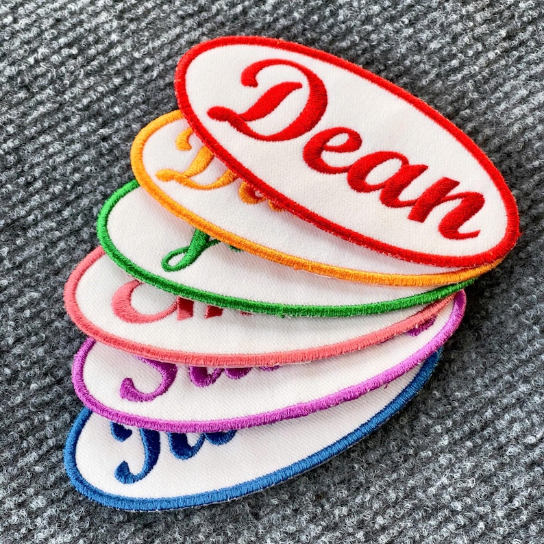 May include: A stack of five oval-shaped embroidered patches with white fabric and colorful borders. The top patch is red with the name "Dean" embroidered in white. The patches are stacked in a rainbow order from top to bottom: red, orange, green, pink, and blue.