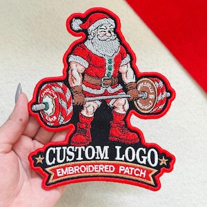 May include: An embroidered patch featuring a muscular Santa Claus lifting a barbell with red and white striped weights. The patch has a red border and a black banner at the bottom with the white text CUSTOM LOGO EMBROIDERED PATCH.