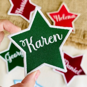 May include: A green star-shaped embroidered patch with the name "Karen" in white script. Other star patches in red and green are visible, each with a different name. The patches have a white border.