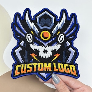 May include: An embroidered patch featuring a stylized skull-like mascot design in shades of blue, white, and black, with a central orange circle. The text CUSTOM LOGO is stitched in bold yellow and orange letters at the bottom of the emblem.