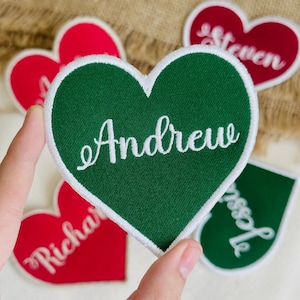 May include: A green heart-shaped patch with the name "Andrew" embroidered in white cursive. Other heart-shaped patches in red and green are visible, each with a different name. The patches have a white border and are likely for personalization.