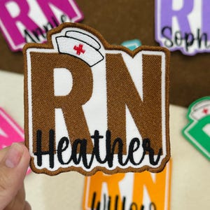 May include: A brown and white embroidered patch with the letters "RN" and the name "Heather" in black script. A nurse's cap with a red cross is above the letters. Other patches are visible in the background.