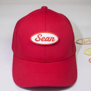 May include: Red baseball cap with a white oval patch that says "Sean" in red embroidered letters.
