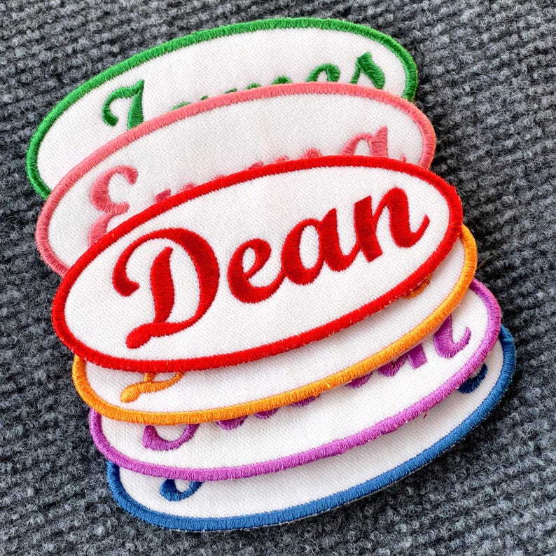 May include: A stack of five oval-shaped embroidered patches with white backgrounds and colorful borders. The top patch is green with the name "James" embroidered in white. The second patch is pink with the name "Emma" embroidered in white. The third patch is red with the name "Dean" embroidered in white. The fourth patch is orange with the name "Olivia" embroidered in white. The bottom patch is blue with the name "Sophie" embroidered in white.