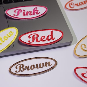 May include: A set of nine white oval fabric patches with different colored embroidered words. The words are "Pink", "Red", "Brown", "Yellow", "Orange", "Green", "Blue", "Black", and "White".