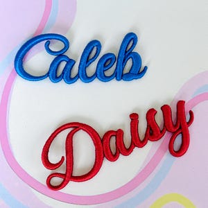 May include: Two embroidered name patches in a cursive script font. The top patch features the name Caleb in a vibrant blue thread, while the bottom patch displays the name Daisy in a bold red thread, both with a raised, textured finish.