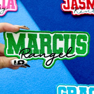 May include: Embroidered patch with the name "MARCUS Rangel" in green and black lettering on a white background. The patch has a green border and is held against a blue background. Other patches are visible.