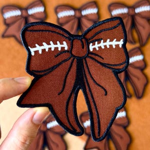 May include: Brown football-themed bow patch with white stitching details. The bow is embroidered with a black outline and has a football design. Several similar patches are visible in the background, suggesting a sports-themed accessory.