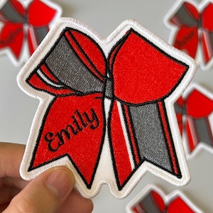 May include: Embroidered red and grey bow patch with the name "Emily" in black script. The bow has a white border, possibly for cheerleading. Other similar patches are visible in the background.