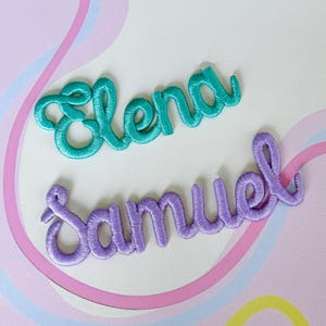 May include: Two embroidered name patches: "Elena" in teal and "Samuel" in purple. The names are in a cursive script style, with a soft, textured appearance. The background is a pastel color scheme with pink and blue accents.