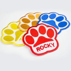 May include: Four paw print patches, one red with the word "ROCKY" in white, one gold, one blue, and one yellow.