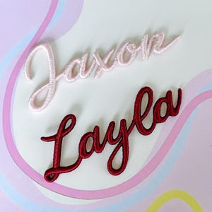 May include: Two embroidered name patches: "Jaxon" in light pink and "Layla" in burgundy. The script lettering is in a cursive style, with a delicate, stitched texture. The background is a soft, pastel design.