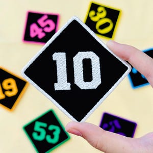 May include: Black diamond-shaped patch with the number 10 in white, surrounded by a white border. Other patches with different numbers in various colors are visible in the background. The patches are likely for clothing or accessories.