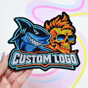 May include: An embroidered patch featuring a blue shark on the left and a yellow and orange skull wearing blue sunglasses on the right. Below the graphics, the text CUSTOM LOGO is stitched in white letters on a light blue and orange background.
