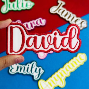 May include: Embroidered name patches in various colors. The patch in focus says "David" in white script with a red border. Other patches include "James", "Julio", "Laura", "Emily", and "Anyname". The patches are on a red and blue background.