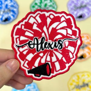 May include: A red and white embroidered patch with the name "Alexis" in black script. The patch features a pom-pom design and a black megaphone. Other patches in blue, green, purple, orange, and yellow are in the background.