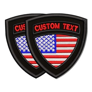 May include: Two black embroidered patches with a shield shape. Each patch features the American flag design with red and white stripes and a blue field of stars. The text "CUSTOM TEXT" is embroidered in red.