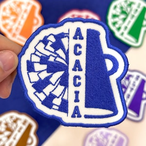May include: A blue and white embroidered patch featuring a megaphone and a pom-pom design. The word "ACACIA" is vertically embroidered in blue on the right side of the patch. The patch has a white border and is likely for a cheerleader.