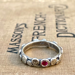May include: A silver ring with a textured band and a single red gemstone. The ring is set on a brown surface.