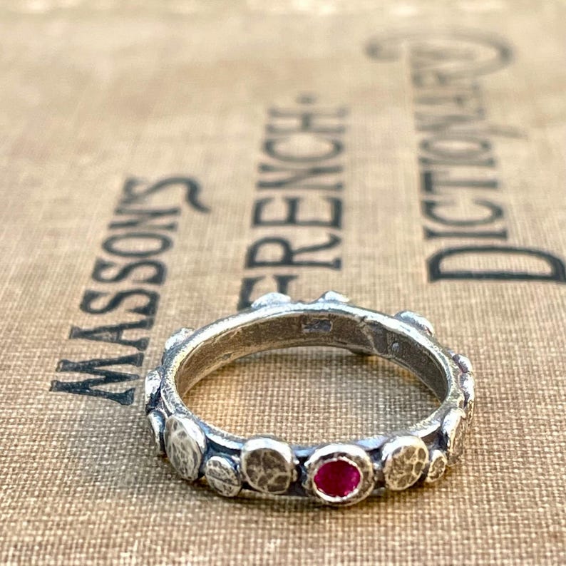 Puede incluir: Un anillo de plata con una banda texturizada y una sola piedra preciosa roja. El anillo est&aacute; colocado sobre una superficie marr&oacute;n.