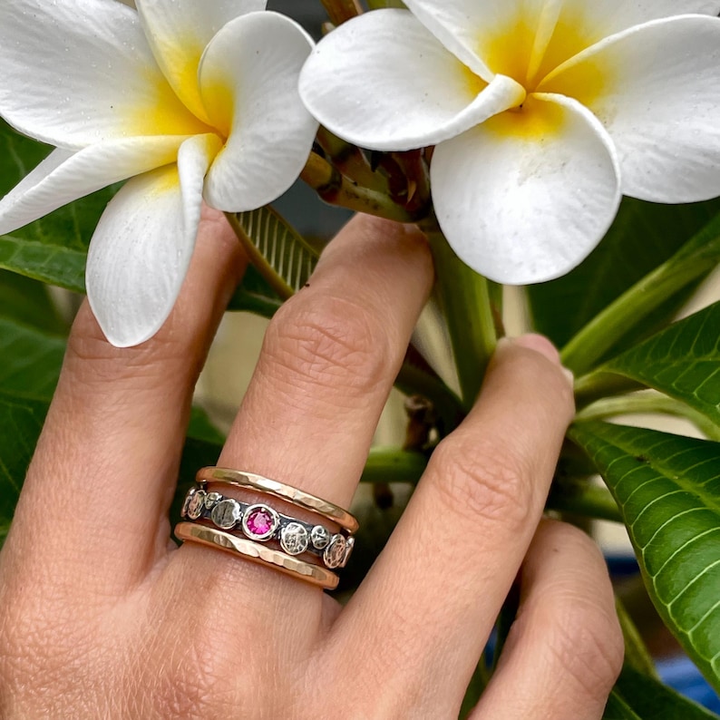 Puede incluir: Un anillo de oro, plata y negro con una piedra preciosa roja. El anillo est&aacute; hecho de tres bandas, cada una con una textura diferente. La banda superior es de oro y tiene un acabado martillado. La banda del medio es de plata y tiene un acabado liso. La banda inferior es negra y tiene un acabado texturizado. La piedra preciosa roja est&aacute; engastada en el centro del anillo.
