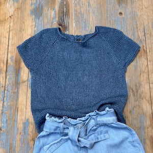 Può includere: Un top blu navy lavorato a maglia con maniche corte e scollo tondo. Abbinato a pantaloncini di jeans azzurri con vita a sacchetto e cintura abbinata. L'outfit è su una superficie di legno rustico.