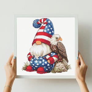May include: A cross-stitch pattern featuring a gnome wearing a tall hat with red and white stripes and blue stars, paired with blue star-patterned overalls and red shoes. A bald eagle sits beside the gnome on a rock, all framed in white.
