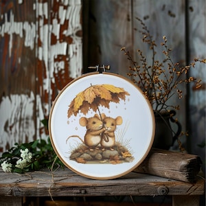 May include: A cross-stitch embroidery hoop featuring two mice sheltering under a leaf umbrella. The artwork is set against a white background, with autumn leaves and a wooden hoop. The color palette includes shades of brown, yellow, and green.