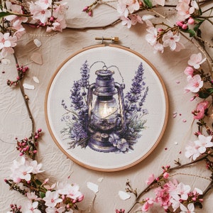 Peut inclure: Une lanterne vintage avec une bougie allumée à l'intérieur, entourée de fleurs de lavande violettes, est présentée dans un cerceau de broderie en bois.
