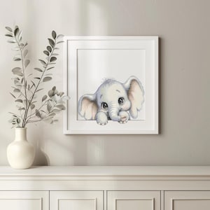 Puede incluir: Una impresión enmarcada con una ilustración en acuarela de un elefante bebé. El elefante es de color gris claro con ojos grandes y expresivos y mira por encima de un borde blanco. La obra de arte está en un marco blanco.