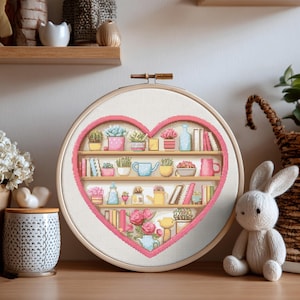 Puede incluir: Un diseño de punto de cruz en forma de corazón que representa una estantería con plantas, libros y objetos decorativos. El diseño está enmarcado en un aro de bordado de madera. Los colores incluyen rosa, verde, amarillo y azul.