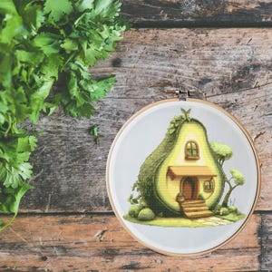 May include: A cross-stitch embroidery hoop featuring a whimsical avocado house design. The avocado is green and yellow, with a small house inside. The design includes a door, windows, and a small tree. The hoop is set against a wooden background.