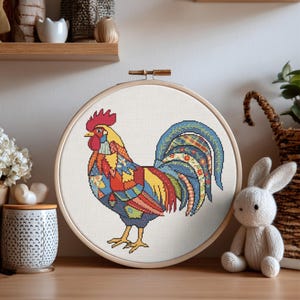 May include: A circular wooden embroidery hoop containing a cross-stitch pattern of a rooster. The rooster features a vibrant patchwork design with red, yellow, blue, and green geometric and floral motifs on a white fabric background.