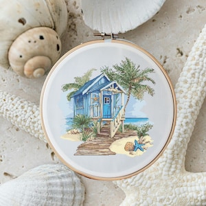 Könnte beinhalten: Eine Kreuzstich-Stickerei, die ein blaues Strandhaus mit einem Holzsteg zum Eingang zeigt. Palmen und ein Seestern befinden sich im Vordergrund.