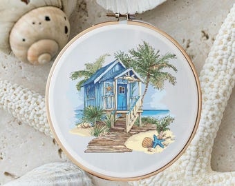 May include: A cross-stitch embroidery of a blue beach house with a wooden walkway leading to the front door. Palm trees and a starfish are in the foreground.