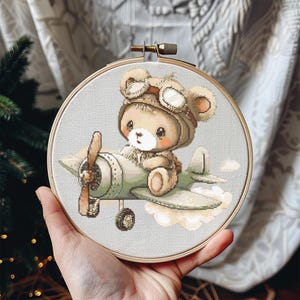 Può includere: Ricamo a punto croce di un orsacchiotto con cappello e occhiali da aviatore, seduto su un aereo d'epoca. Il disegno presenta colori pastello tenui, tra cui beige, marrone e verde chiaro, all'interno di un telaio da ricamo in legno.