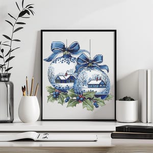 Puede incluir: Obra de arte enmarcada con dos adornos navideños con escenas invernales. Cada adorno está adornado con un lazo azul y un paisaje invernal con casas, árboles y nieve. Acebo y bayas en la parte inferior.